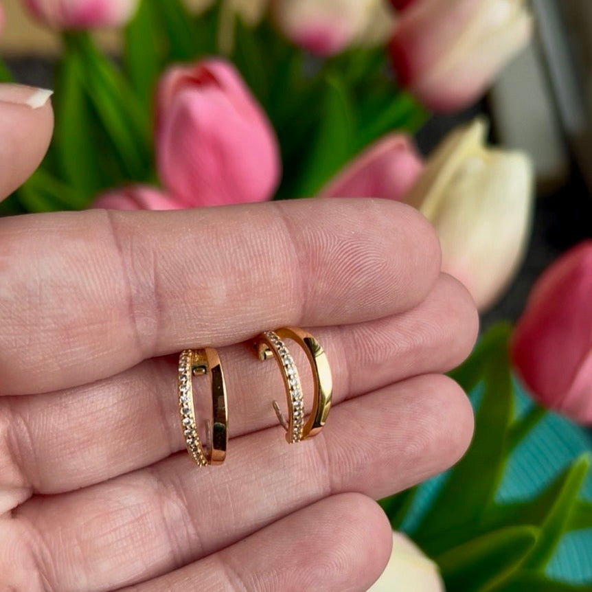 Gold Huggie hoops with sparkling cubic zirconia inlay, perfect for everyday wear. The earrings you reach for on busy mornings. Being held over a flower background.